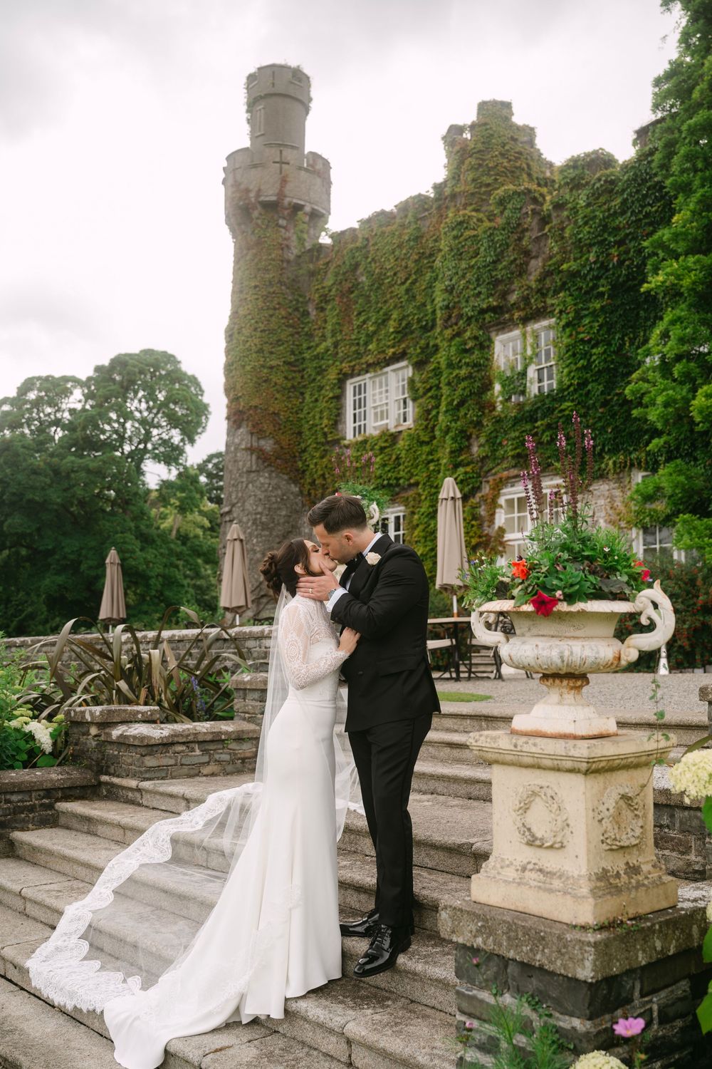 A romantic wedding couple embraces on stone steps outside an ivy-covered castle with a stone tower and ornate urn.