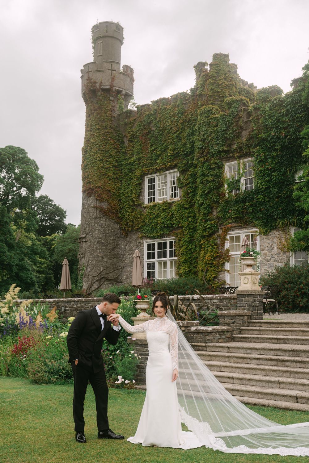 A wedding couple poses for romantic photos in front of an ivy-covered stone castle with grand steps and gardens.