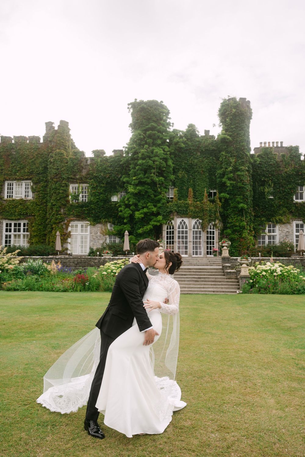 A romantic wedding couple dips and kisses on the lush green lawn of an elegant historic mansion with ivy-covered walls.