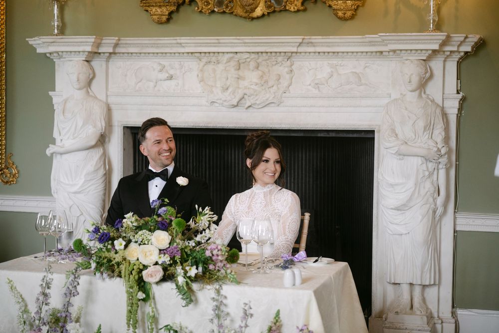 A couple sits at an elegant wedding reception table with marble fireplace and floral arrangements.