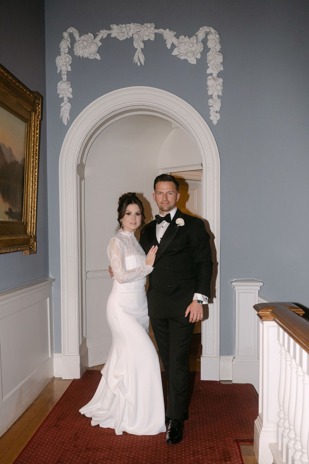 Newlyweds share a romantic moment in a hallway with white archways and red carpeting.