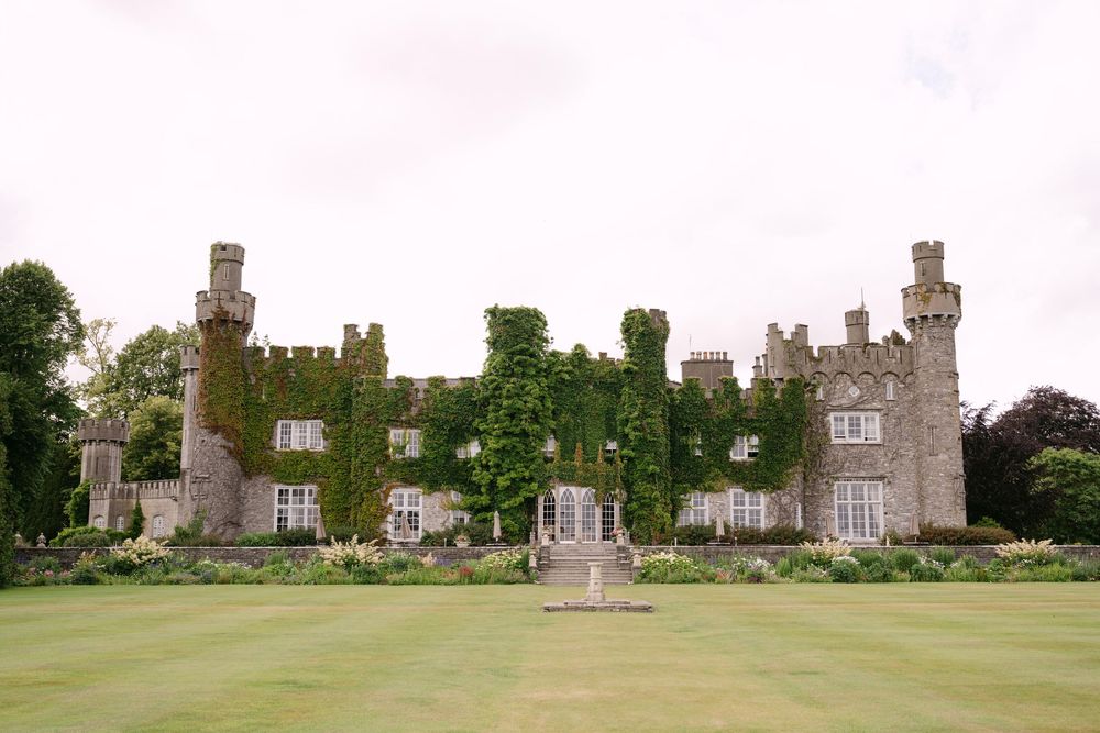 Historic stone castle with ivy-covered walls and turrets stands majestically on a manicured green lawn under cloudy skies.