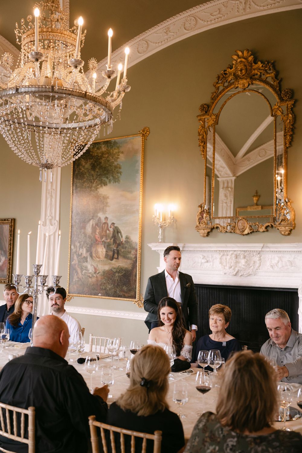 Elegant dining room with crystal chandeliers, ornate mirrors, and wall murals features guests seated at long tables.