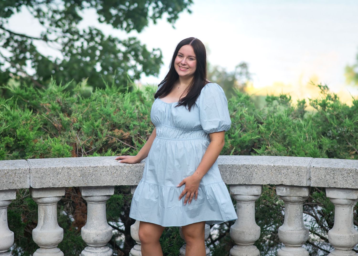 A woman in a light blue denim top poses outdoors against a stone railing with lush greenery in the background.