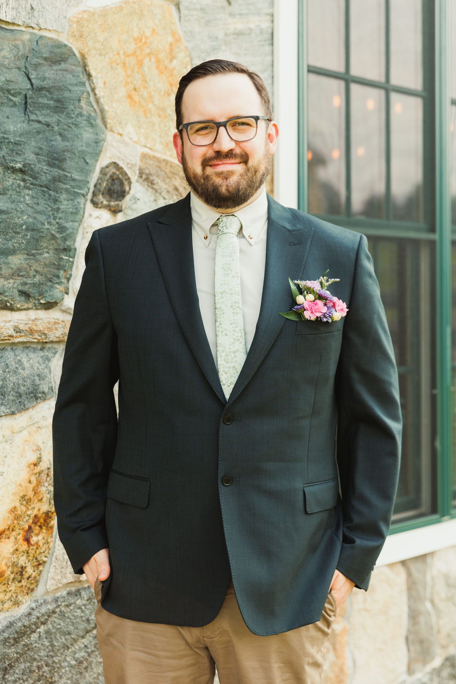 Groom in black blazer and khaki pants poses against stone wall with boutonniere.