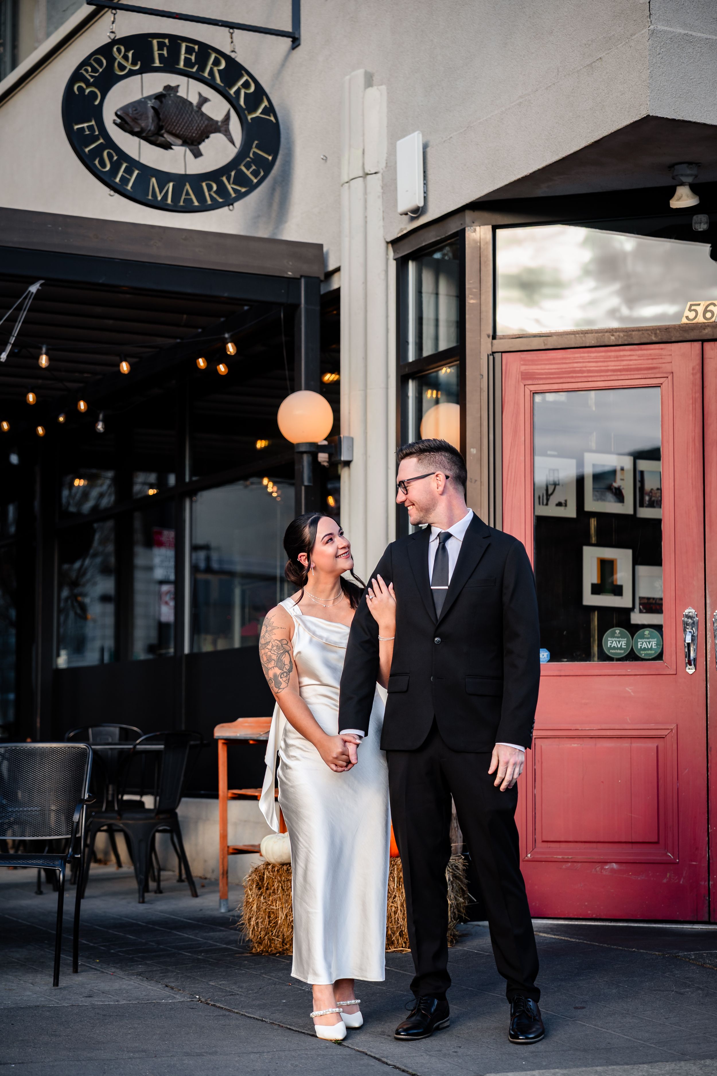 Couple shares playful moments outside a restaurant storefront with red door and string lights.