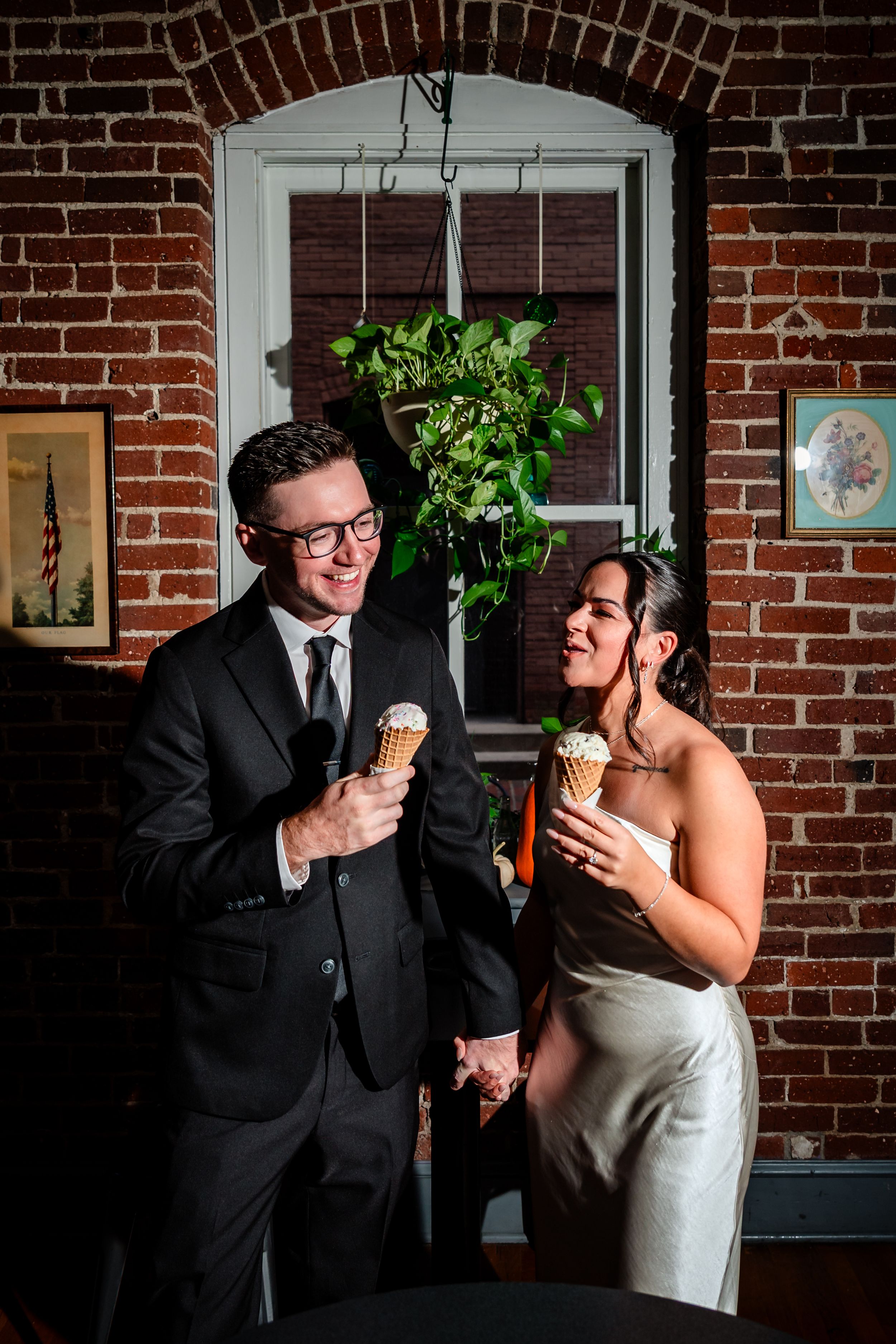 Couple laughs together while posing in front of exposed brick wall with hanging plants.