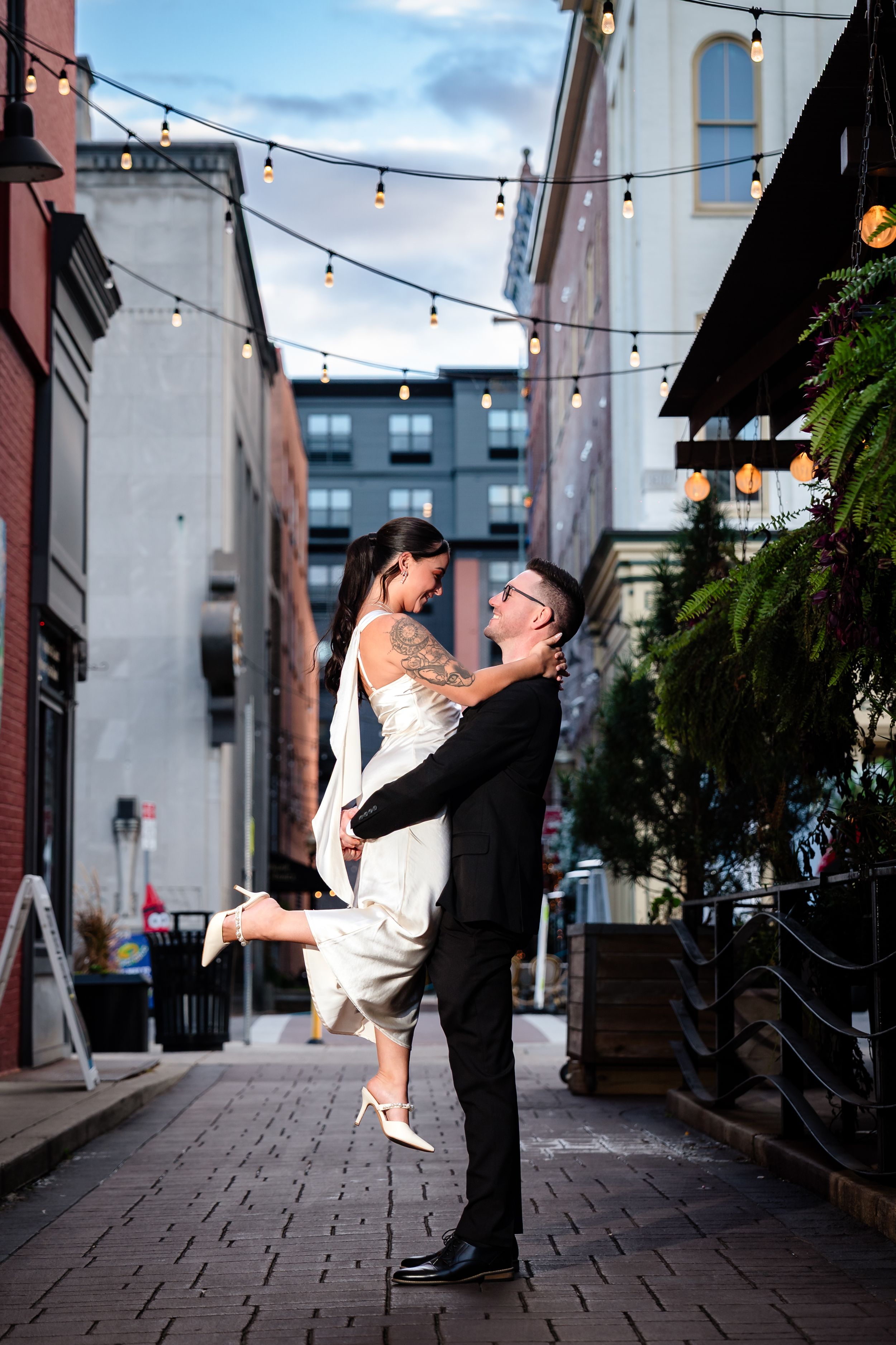 Dancing couple in formal attire beneath string lights in a cobblestone alley.