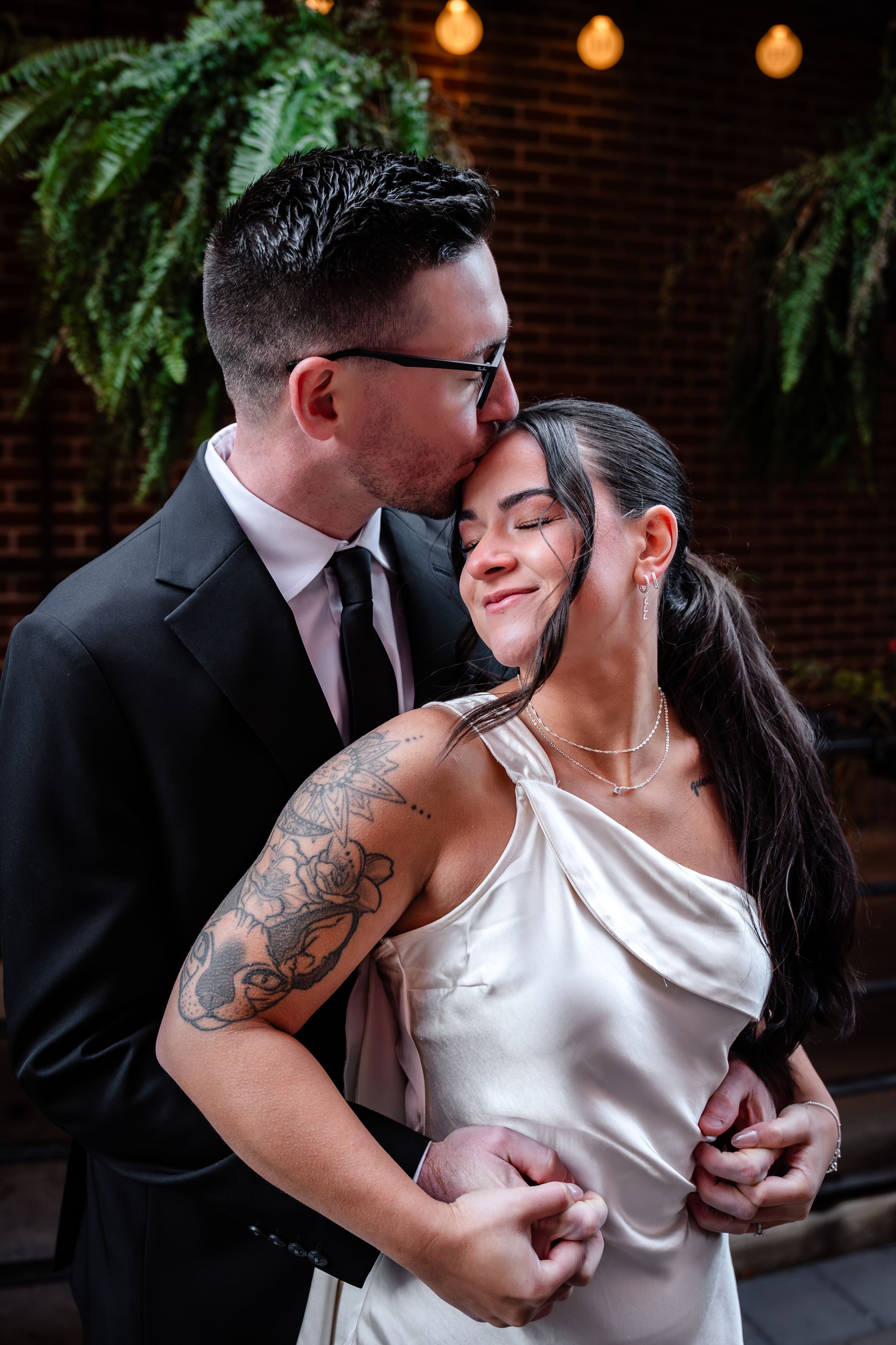 Intimate couple embraces in romantic setting with string lights and greenery.