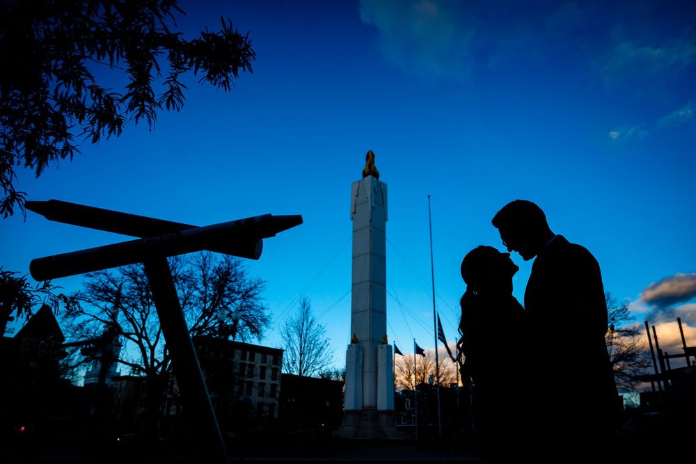 Silhouettes of figures and an airplane against a blue sky with a tall monument in the background at dusk.