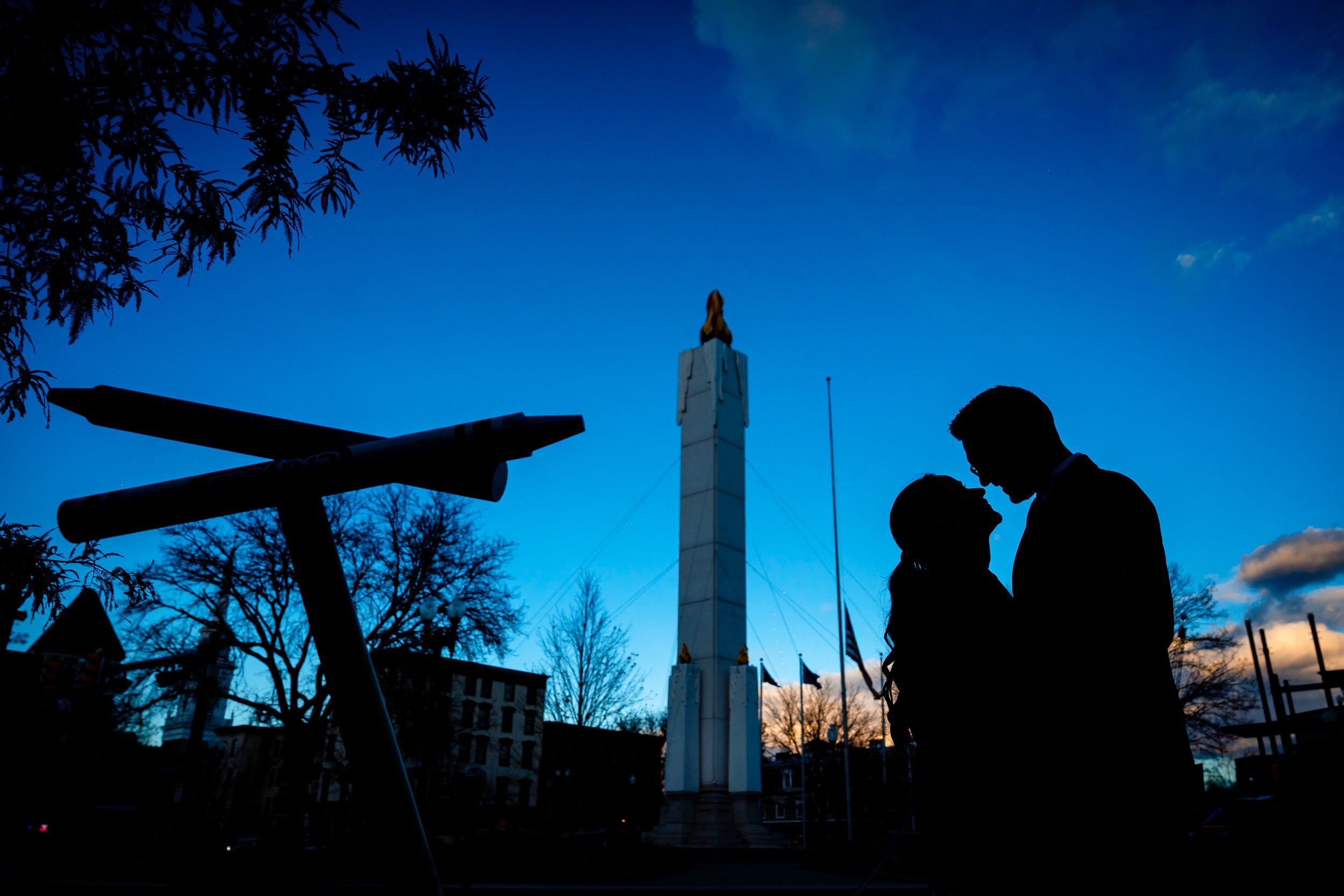 Silhouettes of figures and an airplane against a blue sky with a tall monument in the background at dusk.