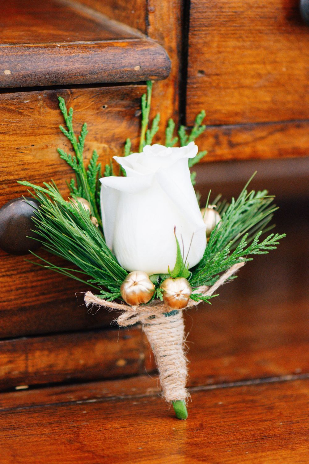 White rose boutonnieres with greenery and twine wrap displayed on wooden surface.