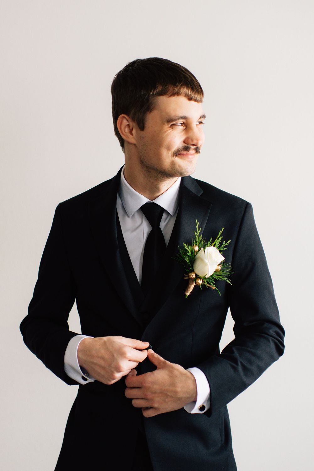 Person in dark suit and white boutonniere making expressive gestures against white wall.
