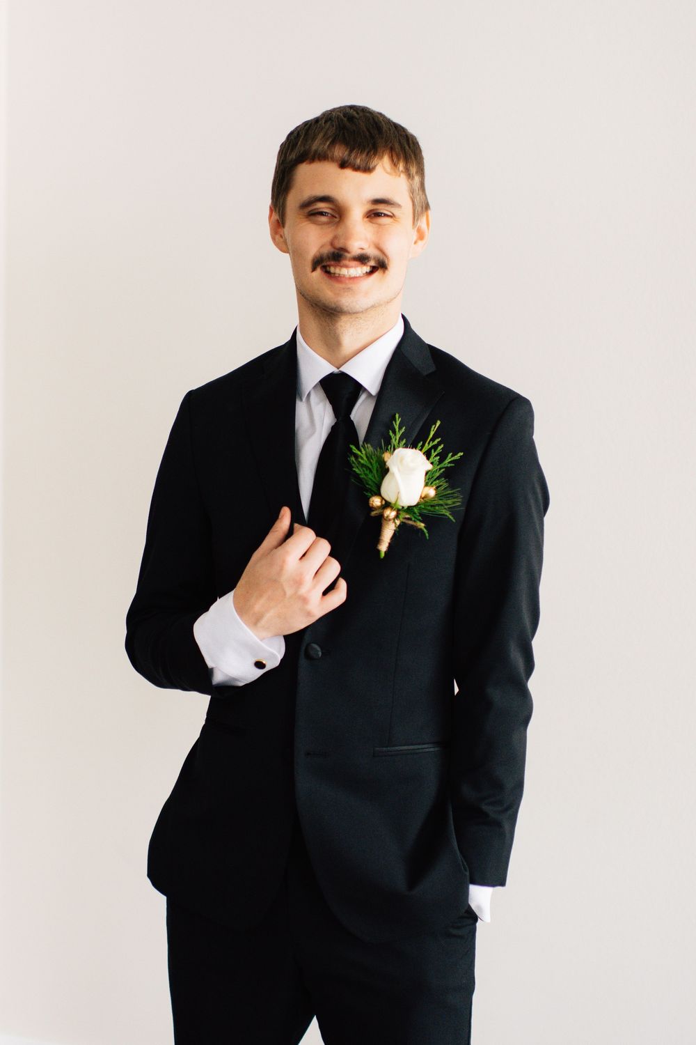 Groomsman in classic black tuxedo with white boutonniere poses against white backdrop in studio portraits.