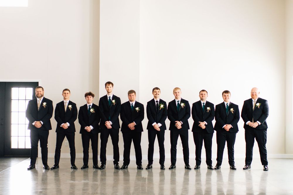 Wedding party in black suits lined up against white wall for creative group photo.
