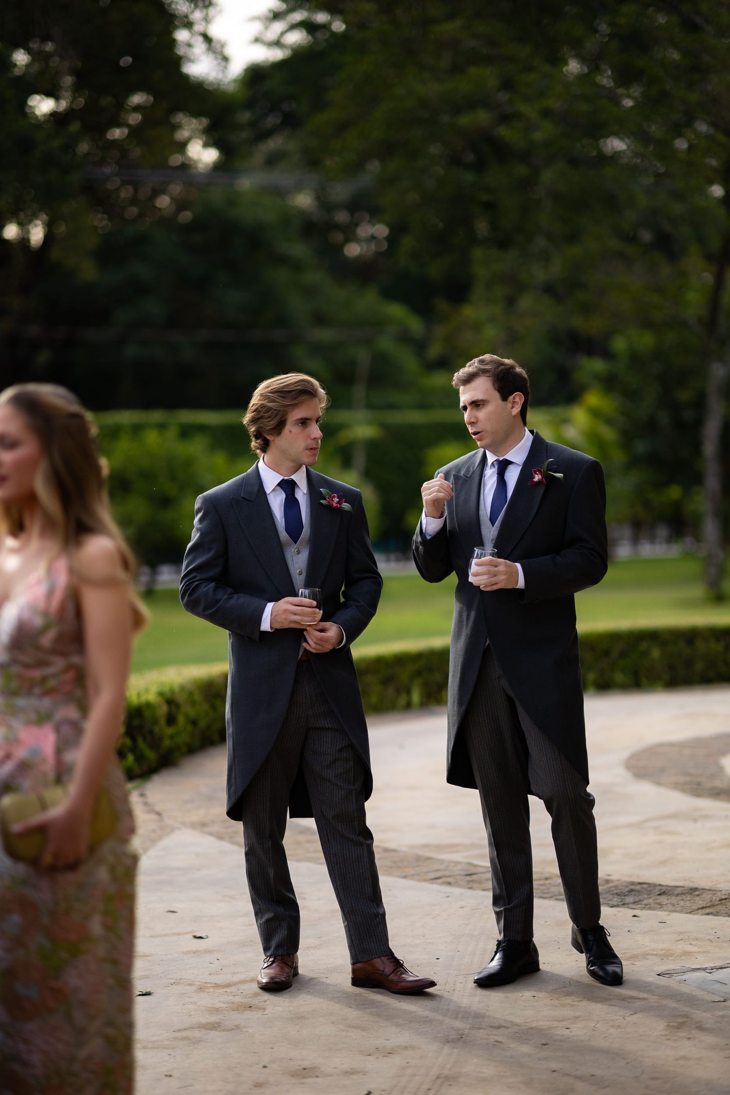 Groomsmen in dark suits stand casually on stone path during outdoor wedding photo session.