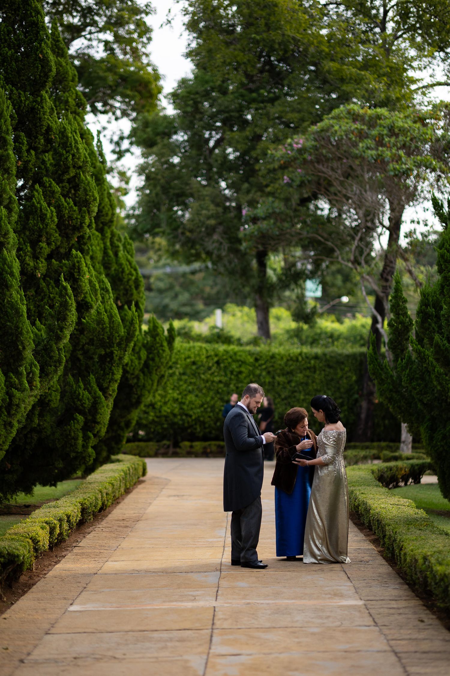 A small ceremony takes place on a garden path lined with manicured hedges and cypress trees on a peaceful afternoon.