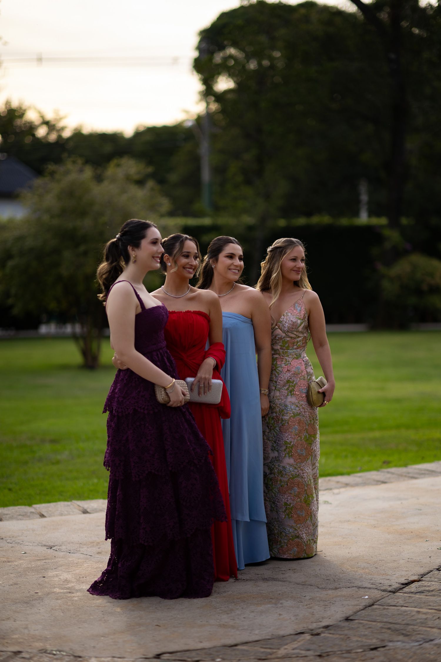 A group in formal dresses sits together on outdoor steps at sunset during an elegant event.