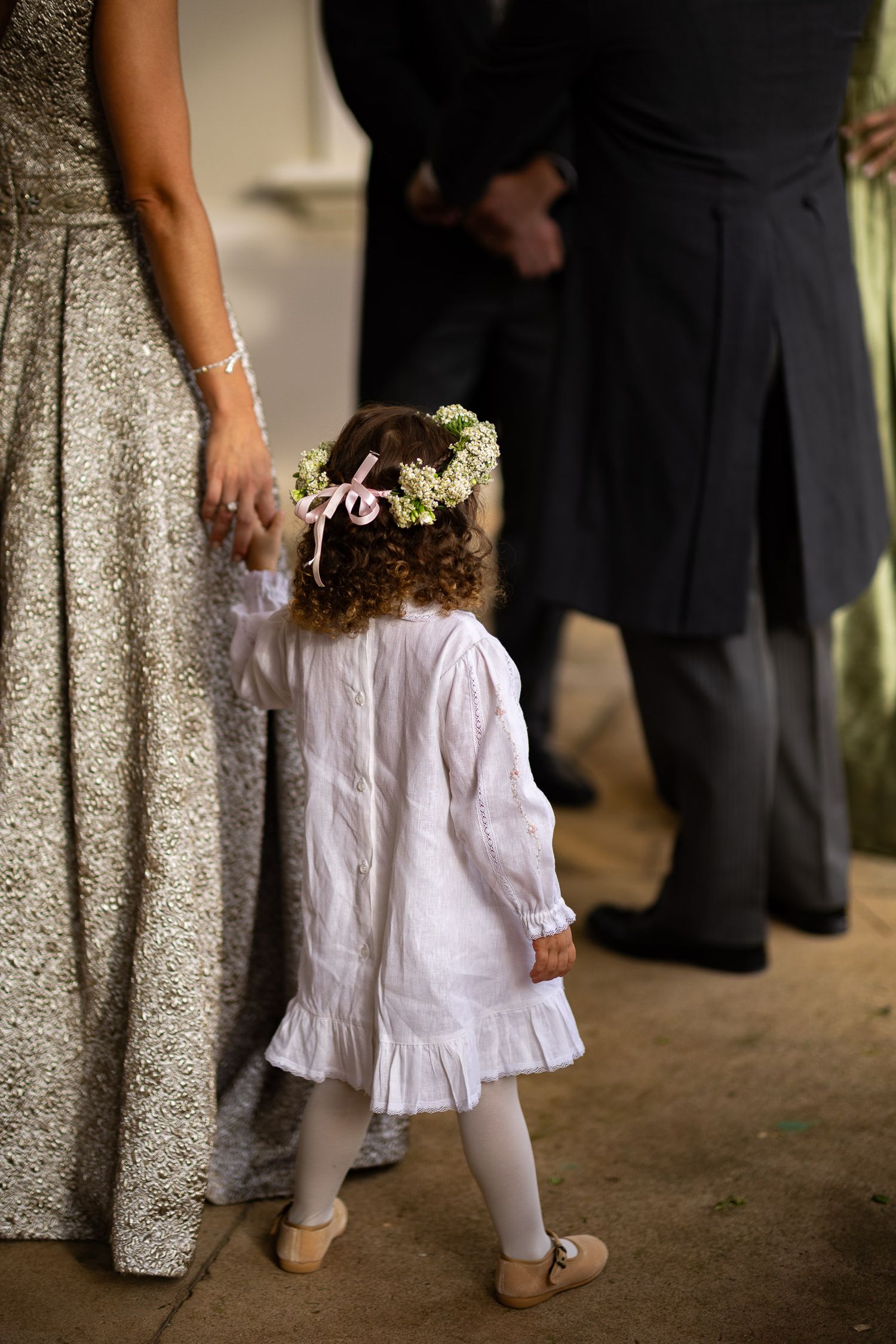 A small child in a white dress holds a flower basket while walking at a formal event.