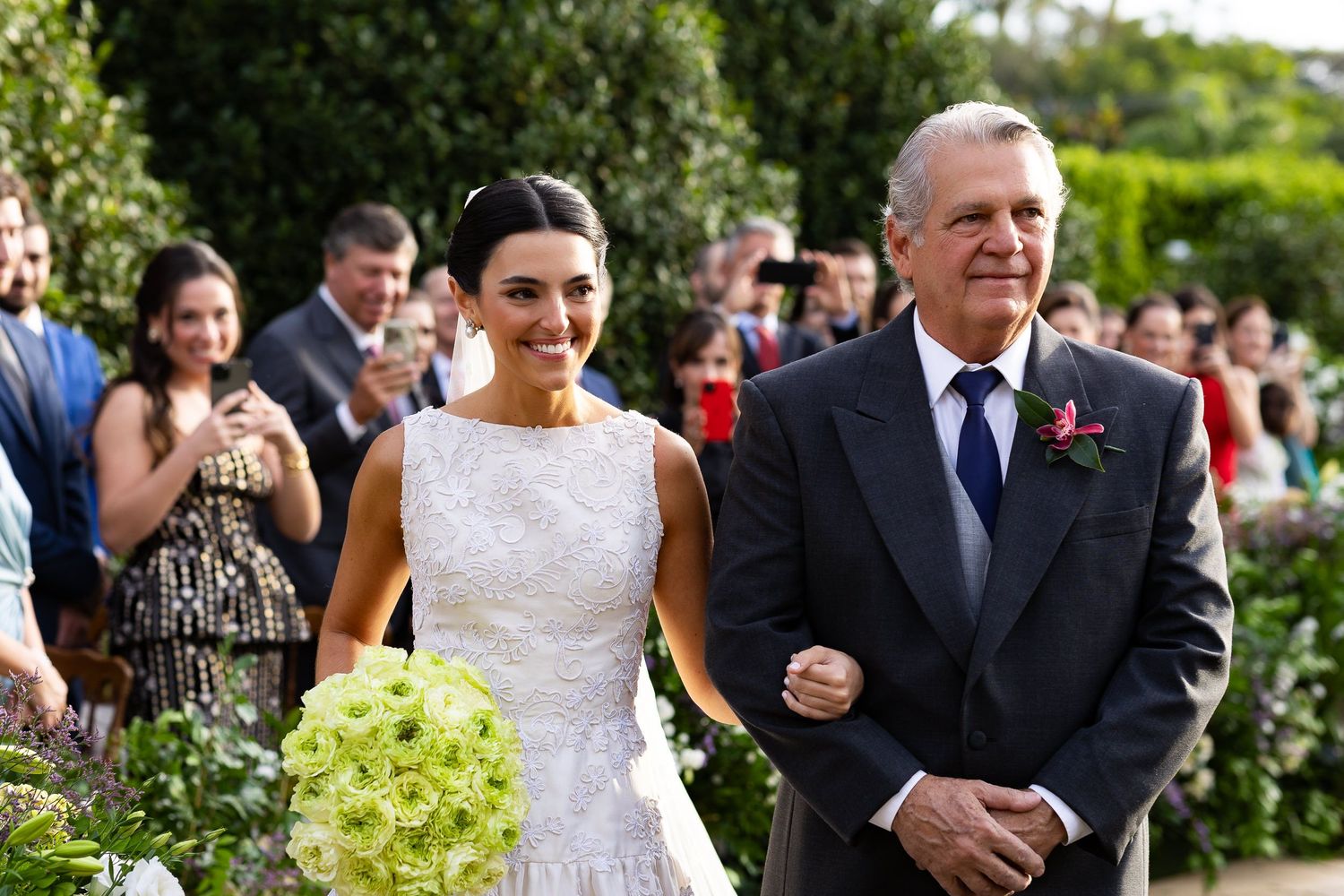 A bride and escort walk down an outdoor garden path surrounded by wedding guests on a sunny day.