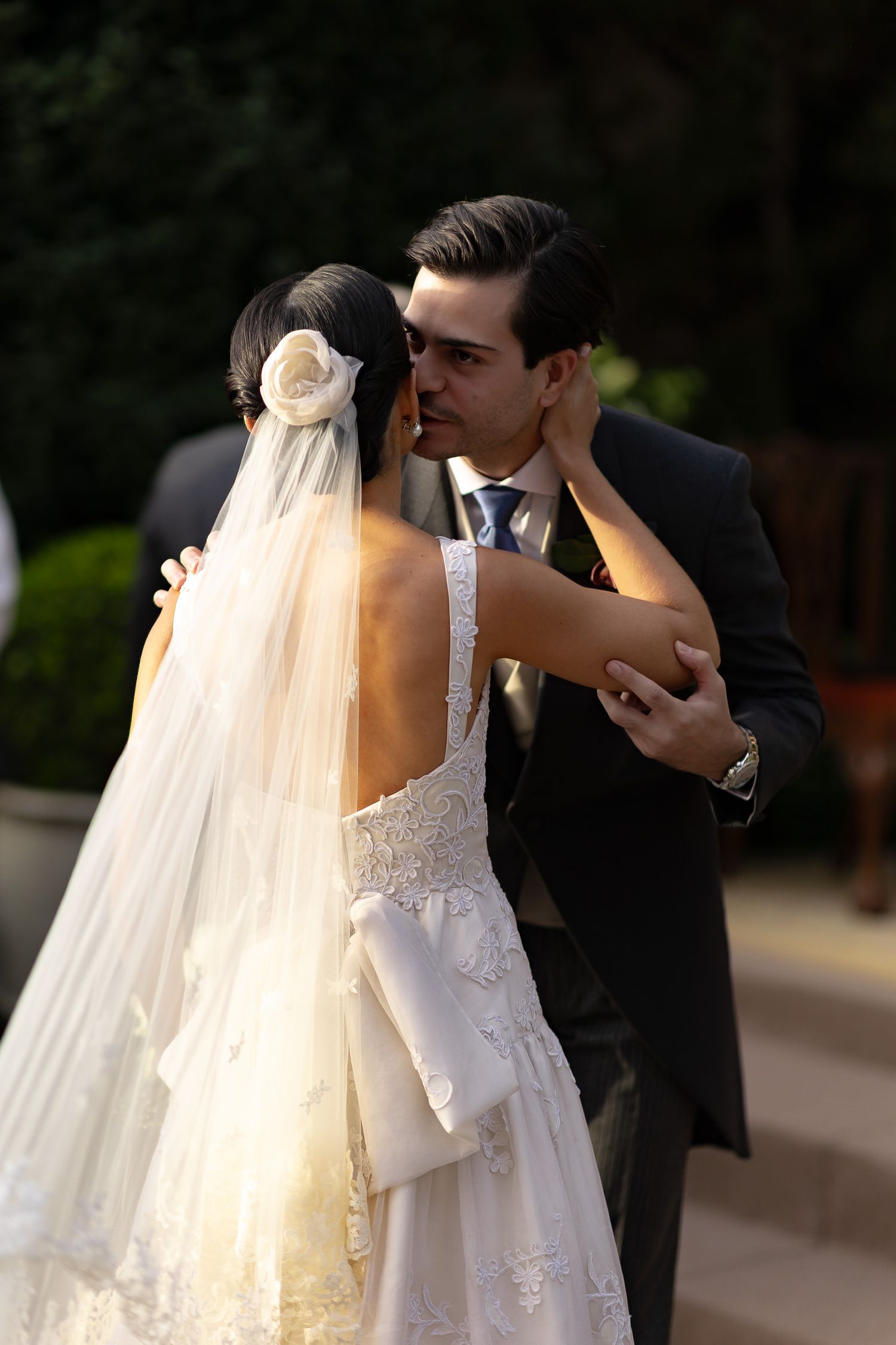 A bride and groom share a romantic kiss outdoors while her long white veil flows in the sunlight.