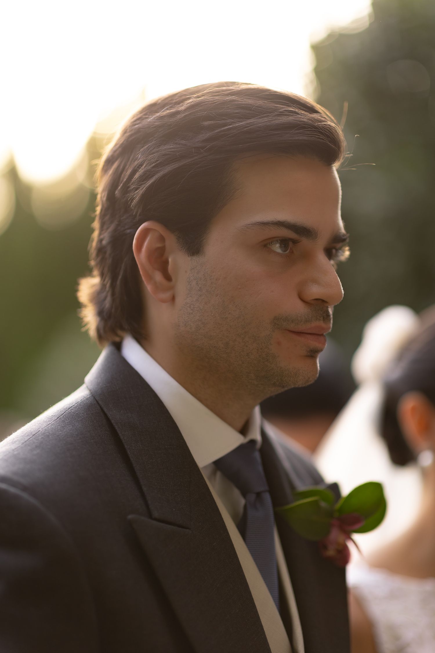 A man in a dark suit and blue tie is shown in profile view at an outdoor event during golden hour.