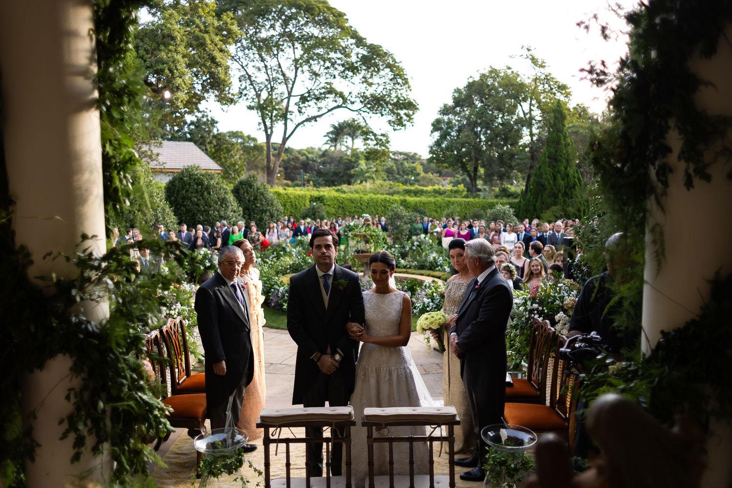 Wedding ceremony taking place in a lush garden setting with guests gathered under trees and decorative columns.
