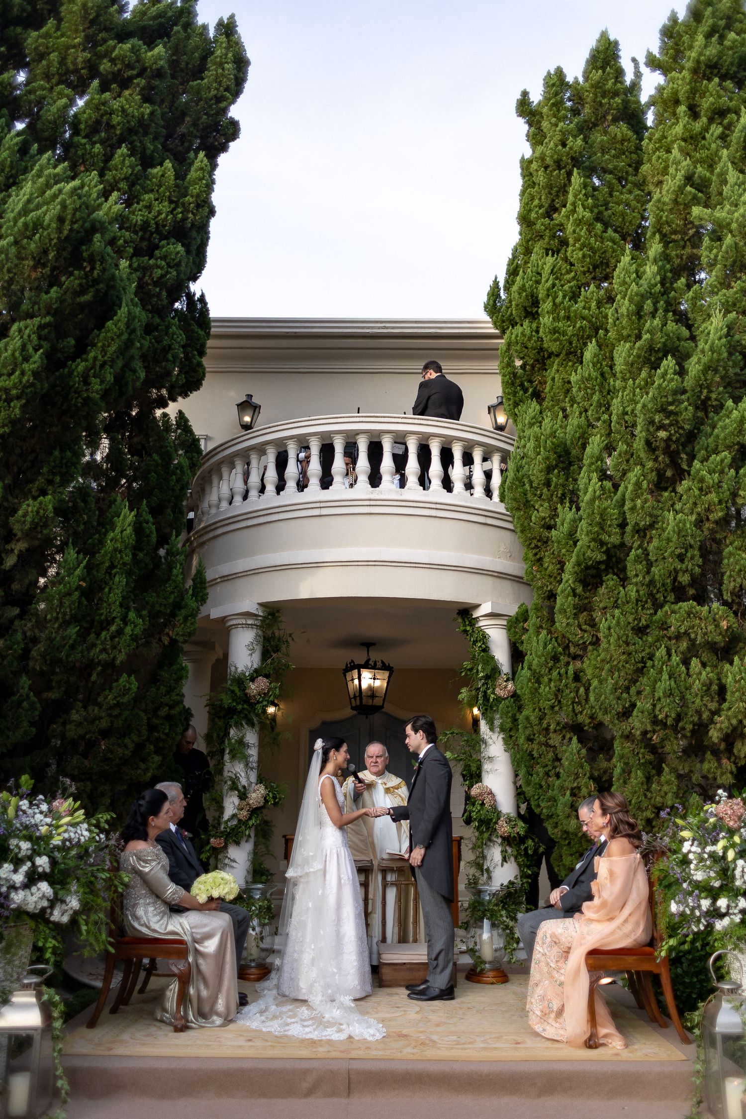 Wedding ceremony taking place under an elegant white archway surrounded by lush cypress trees and garden landscaping.