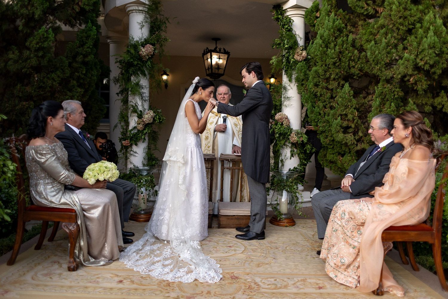 An intimate wedding ceremony takes place in a garden courtyard with white pillars and lanterns.