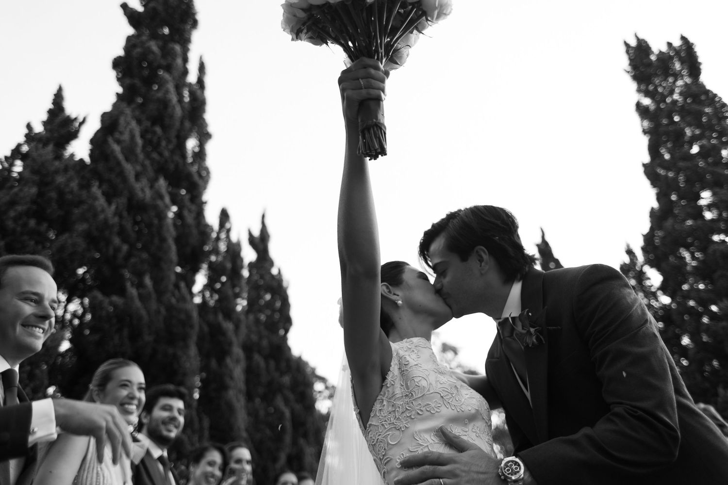 A joyous outdoor wedding celebration captured in black and white with tall cypress trees in the background.
