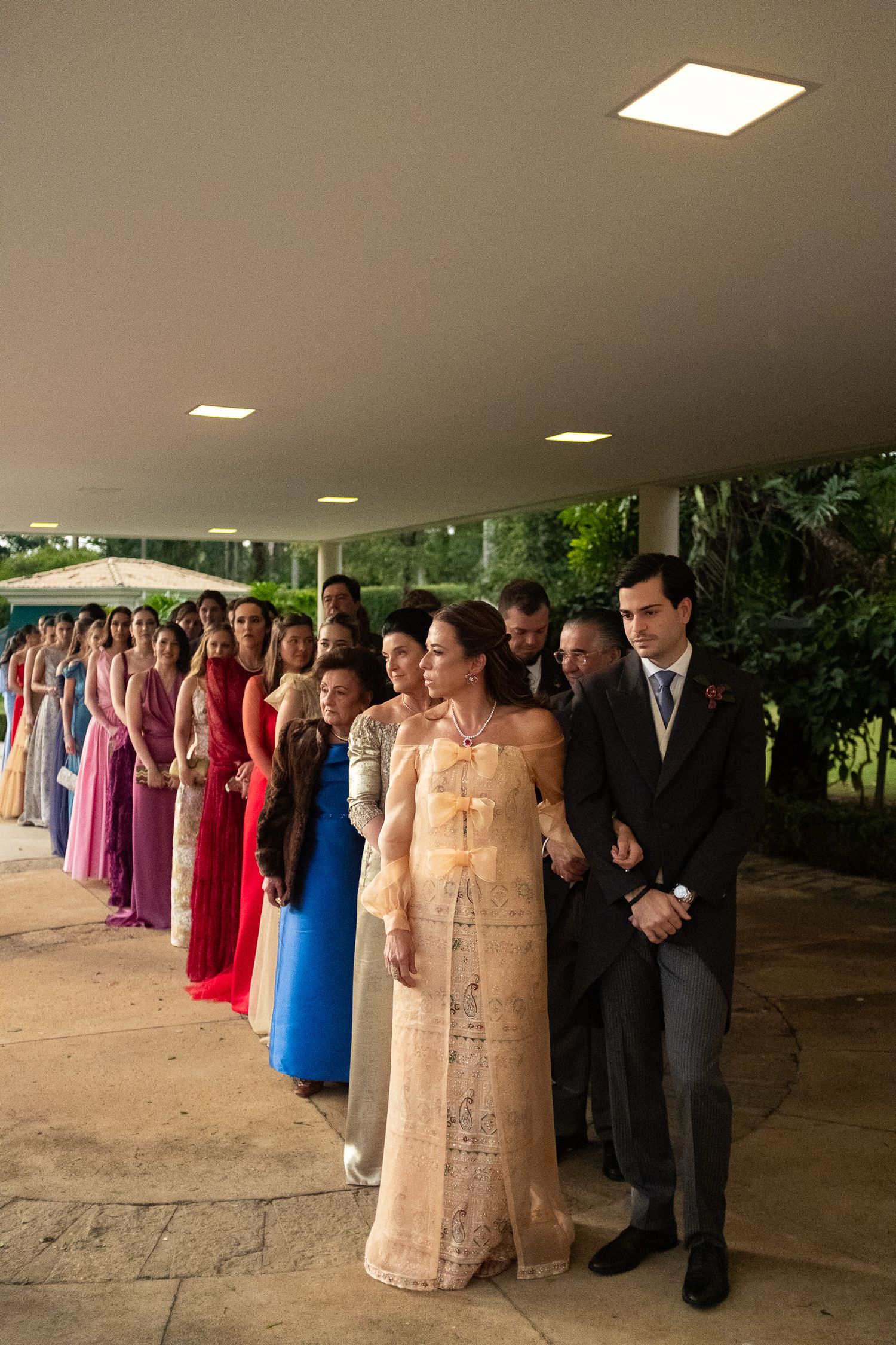 A formal event with attendees lined up in colorful evening gowns and suits under a covered outdoor area.