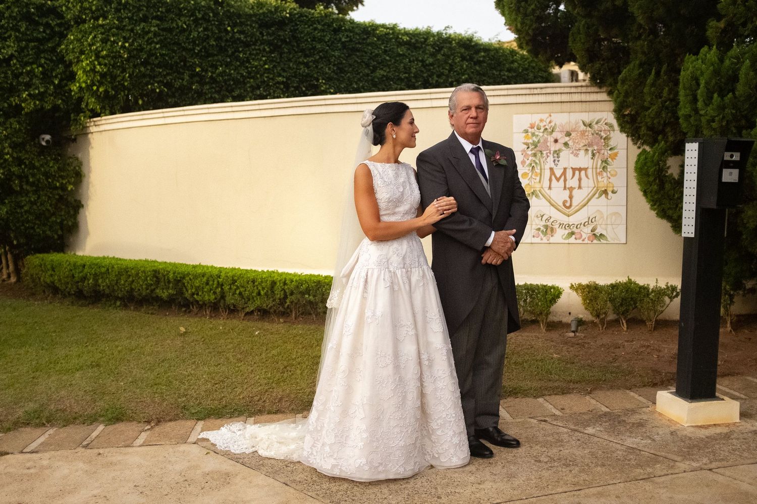 A couple in formal wedding attire stands together on a garden path beside a cream-colored wall with decorative hedges.