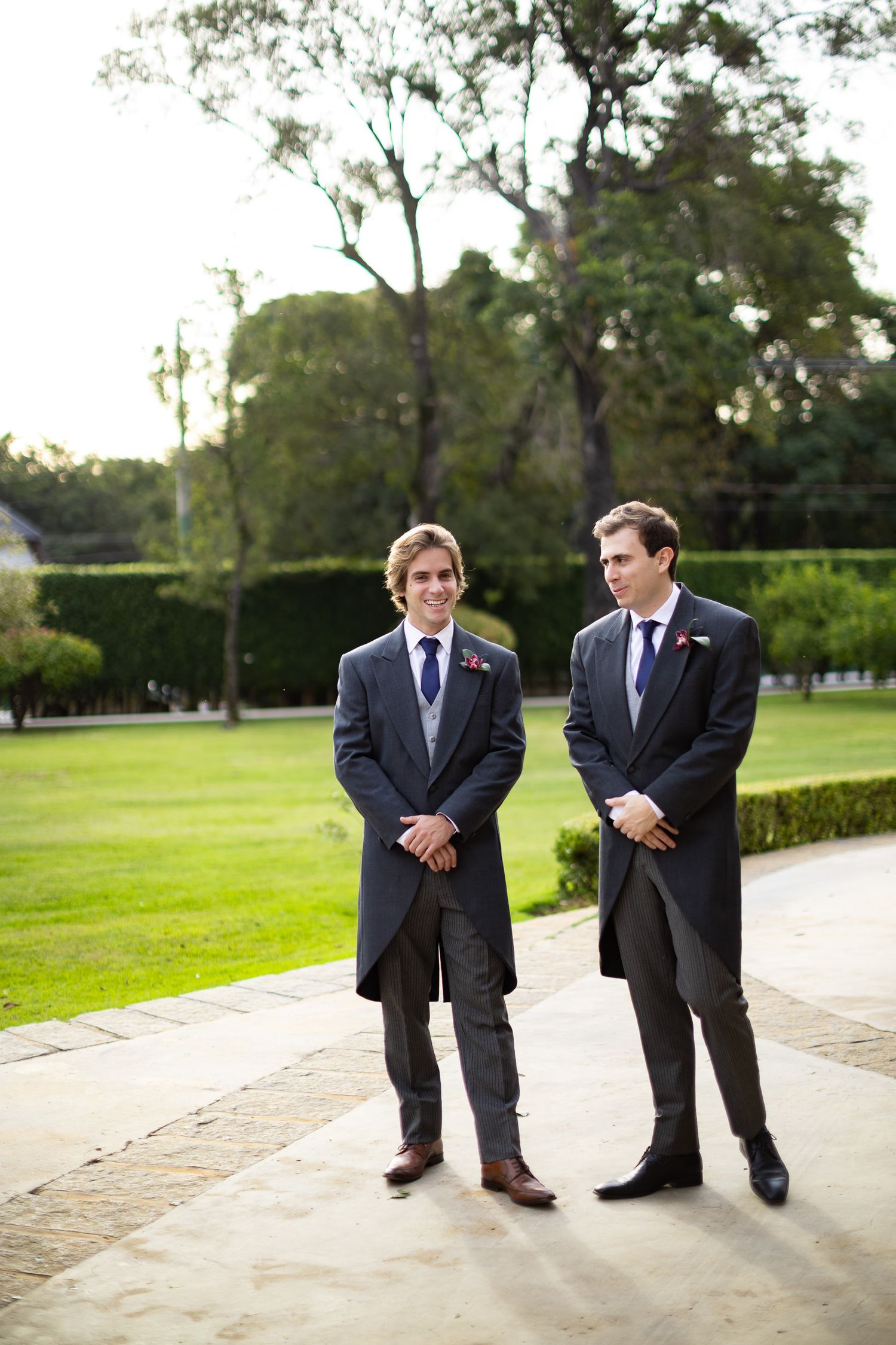 Two groomsmen in dark suits stand together on a pathway with green lawn in background.