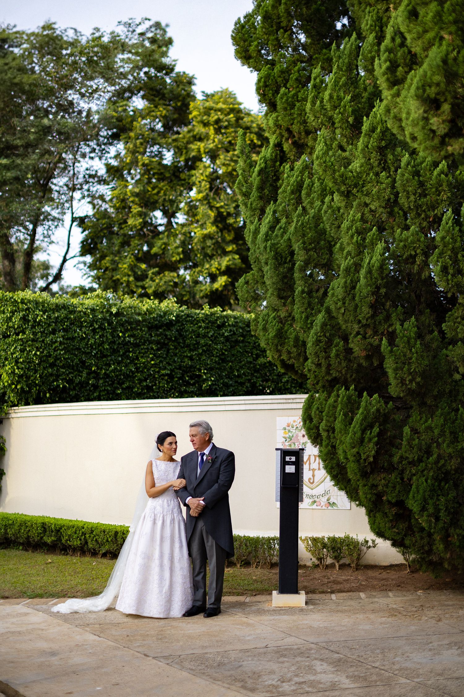 A couple embraces in a garden setting with tall cypress trees and white walls on their wedding day.