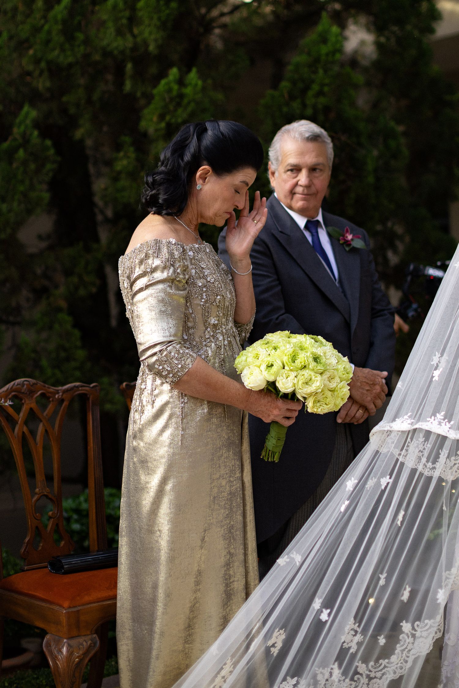 A couple in formal attire stands together at an outdoor ceremony with yellow bouquet and wooden chairs visible.