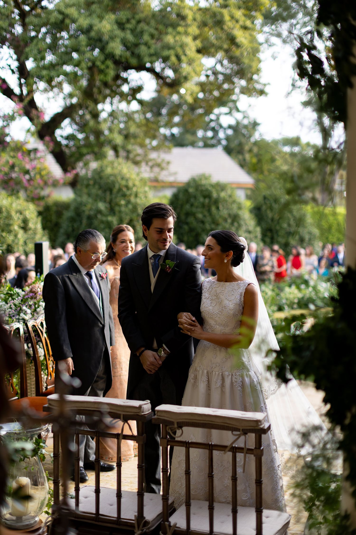 A group of wedding guests gather and chat together on a shaded outdoor patio during a daytime celebration.
