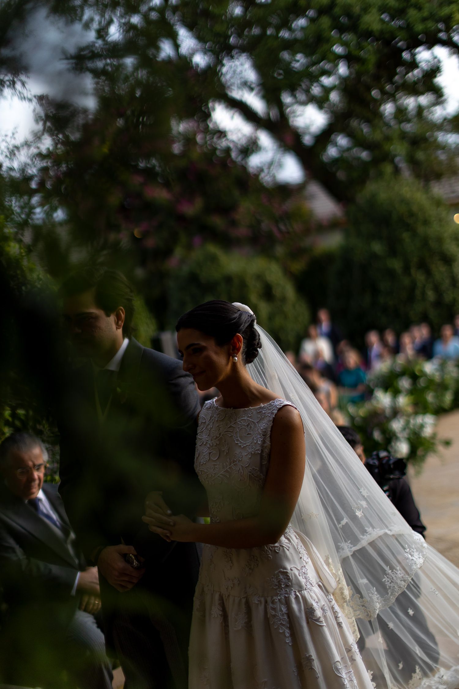 A bride in a white lace wedding dress and flowing veil stands outdoors during a garden ceremony.