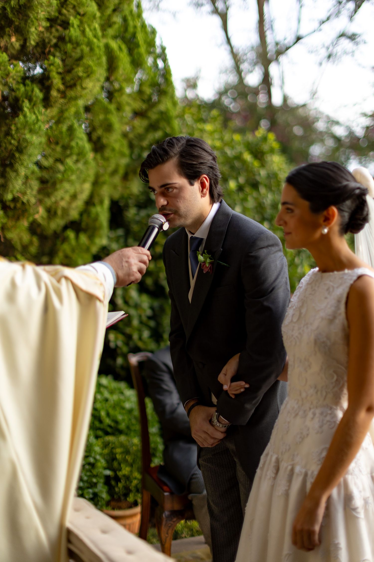 A couple stands at an outdoor wedding ceremony while a priest in cream robes holds a microphone during the service.