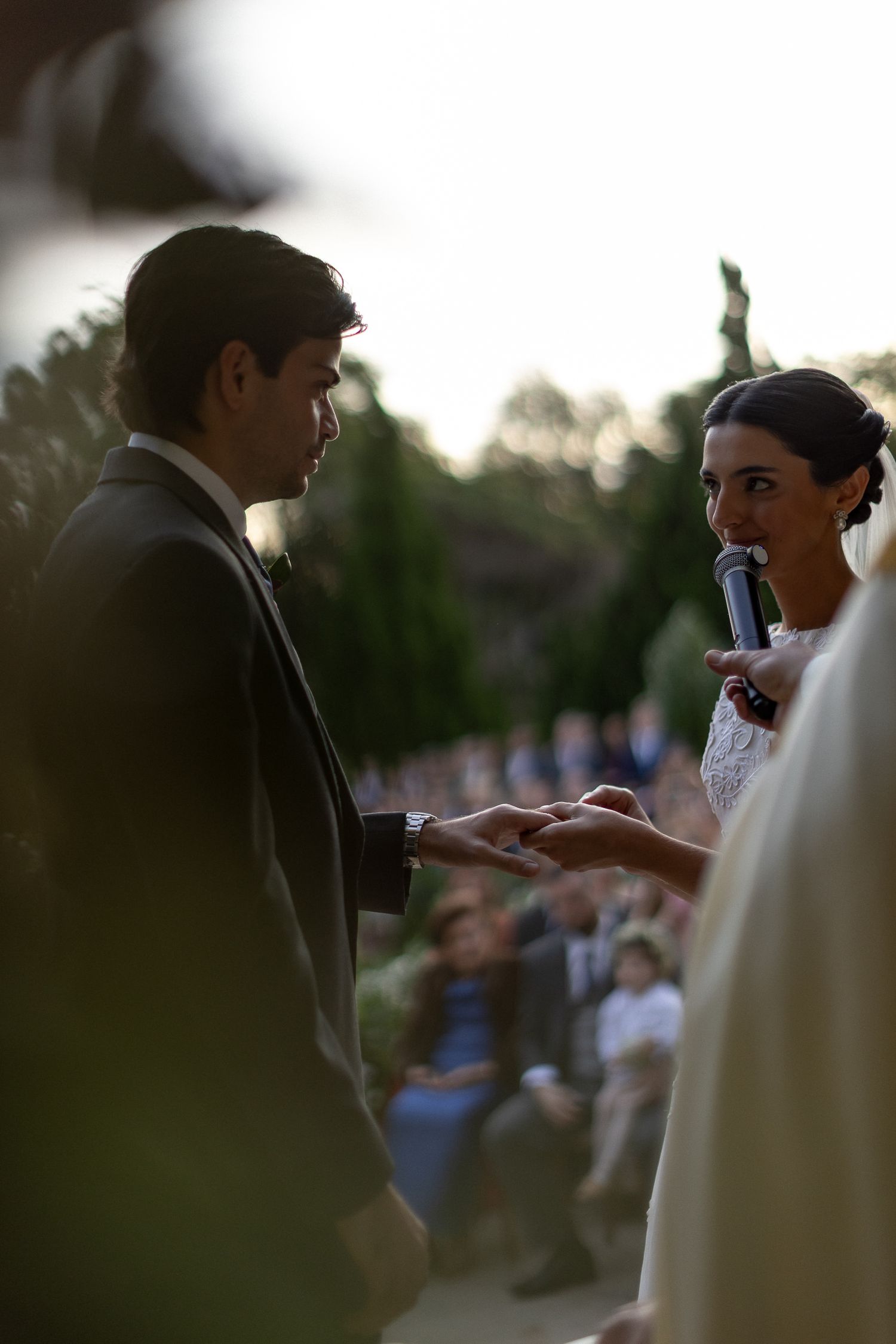 A couple exchanges rings during an outdoor wedding ceremony at sunset with guests in the background.