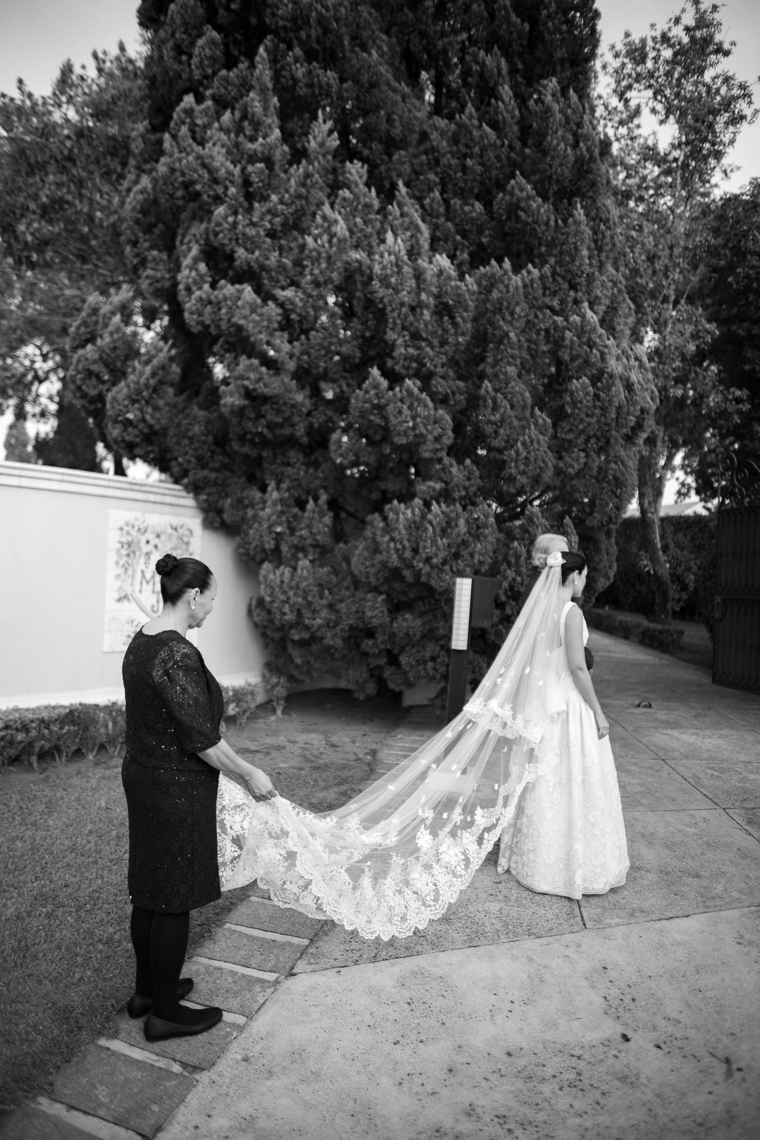 A bride in a flowing white gown with long lace train stands on steps while an attendant adjusts the veil.