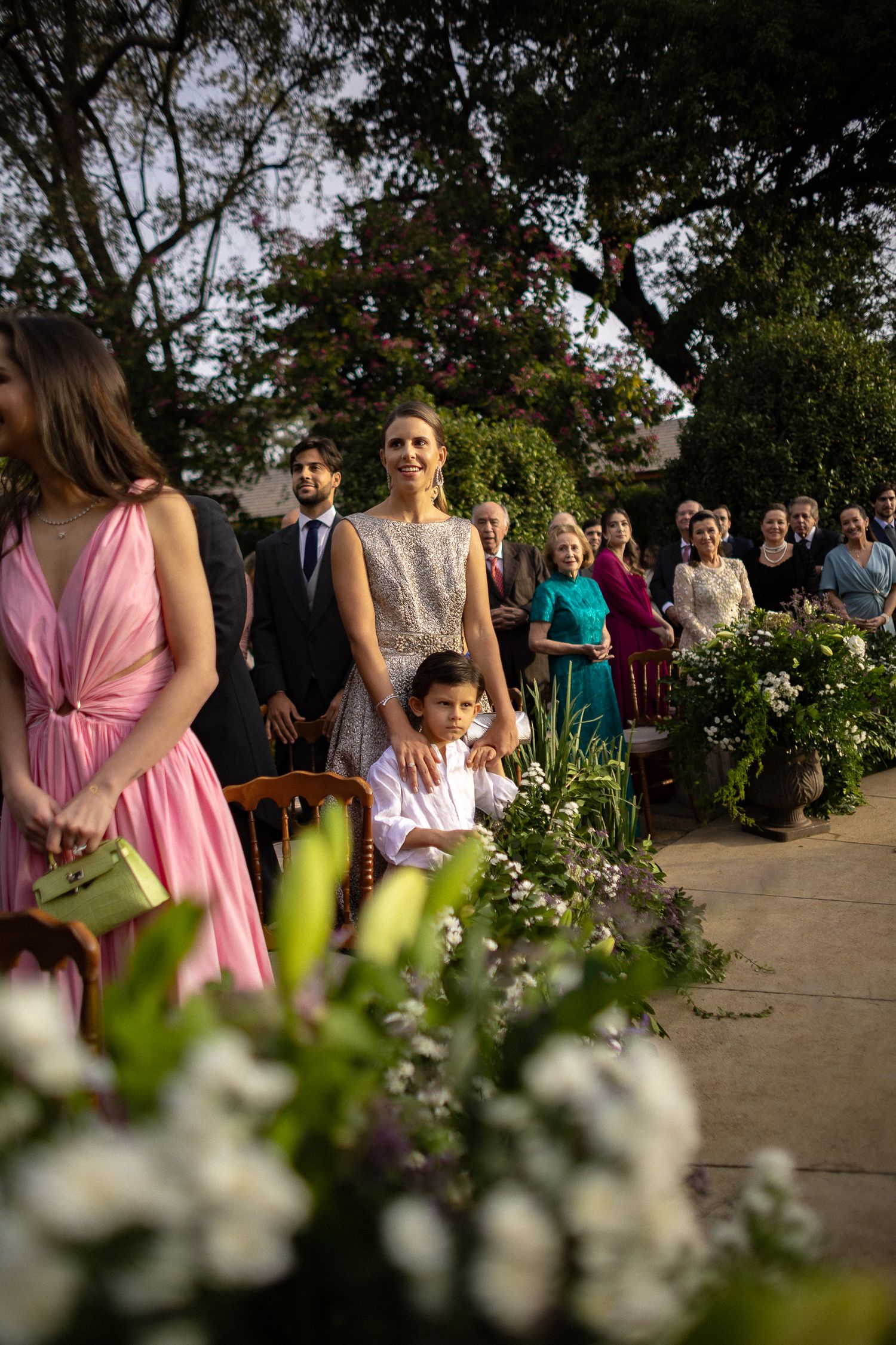 Wedding guests gather outdoors for a ceremony with white flowers and greenery in the foreground.