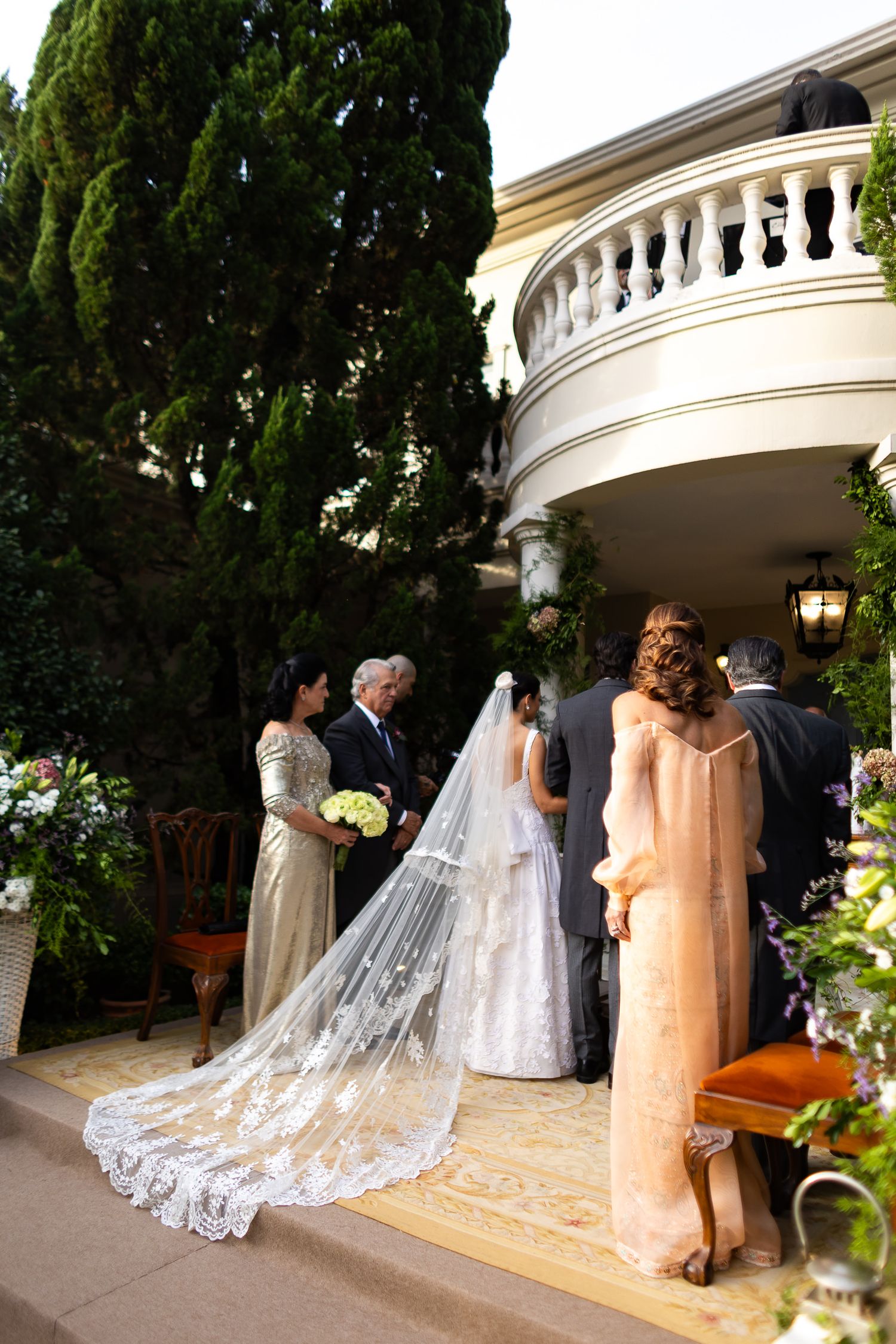 A wedding ceremony procession in an elegant outdoor colonnade with a long flowing white lace veil trailing behind.