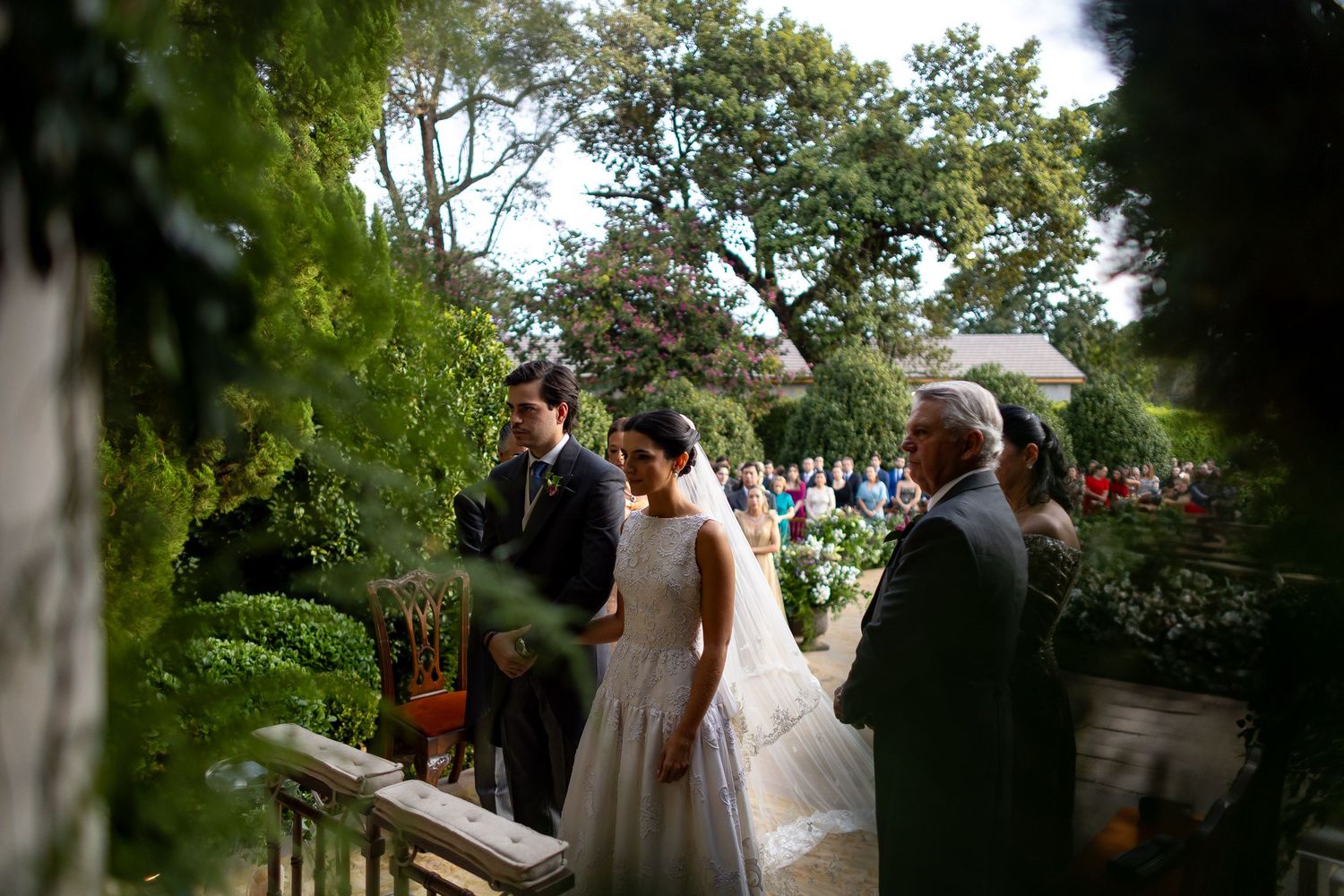 Wedding guests gather outdoors under trees during a ceremony at a scenic garden venue.