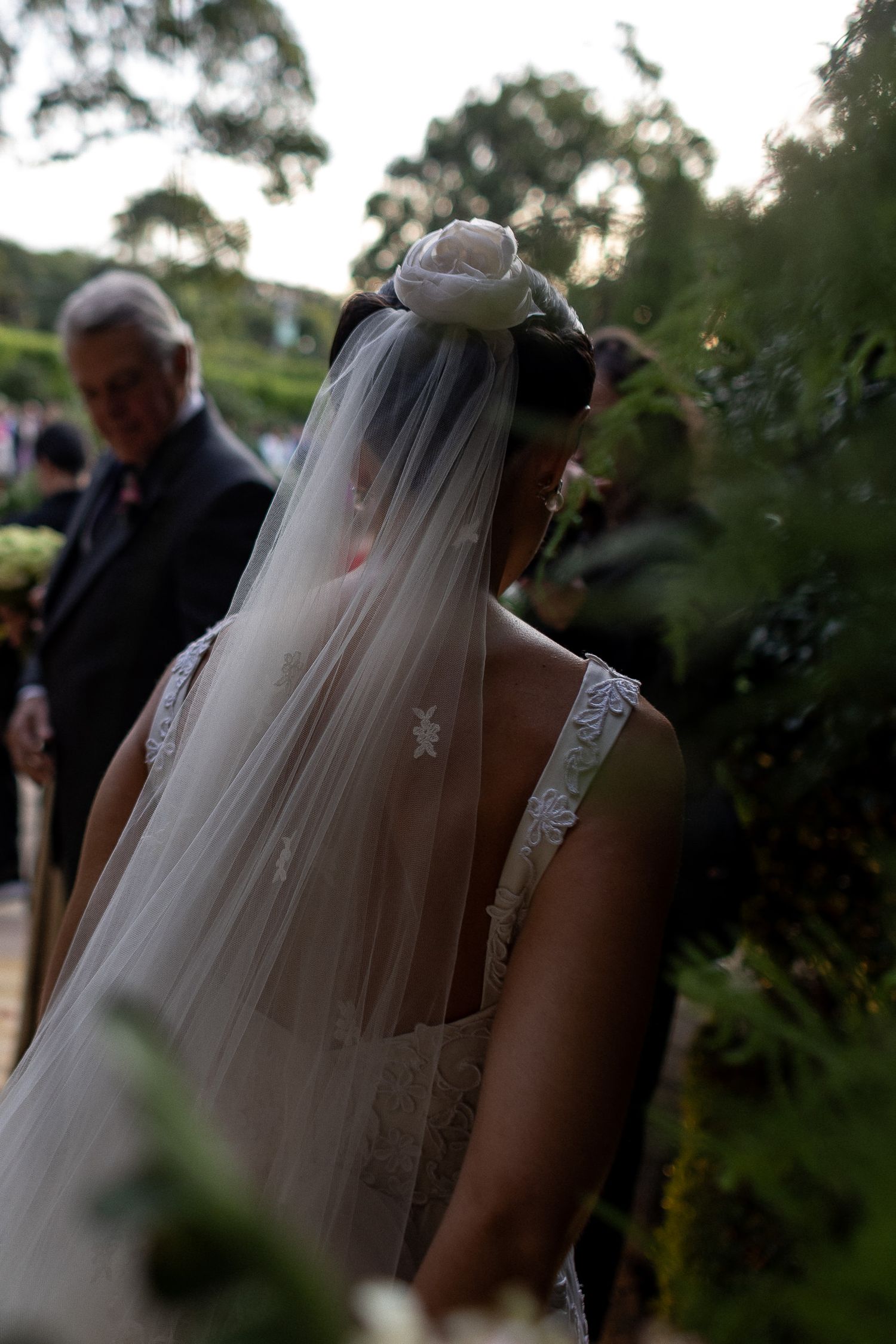A bride in a white wedding dress and long veil stands outdoors during a garden ceremony at dusk.
