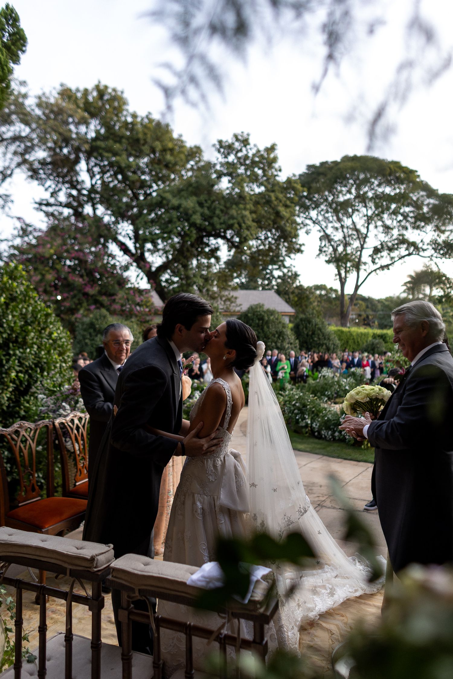 A wedding ceremony takes place outdoors with guests seated on benches under trees during a beautiful autumn day.