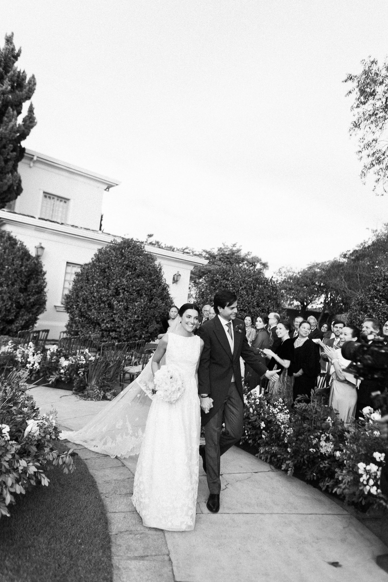 A black and white photograph captures a wedding party walking along a garden path lined with blooming flowers.