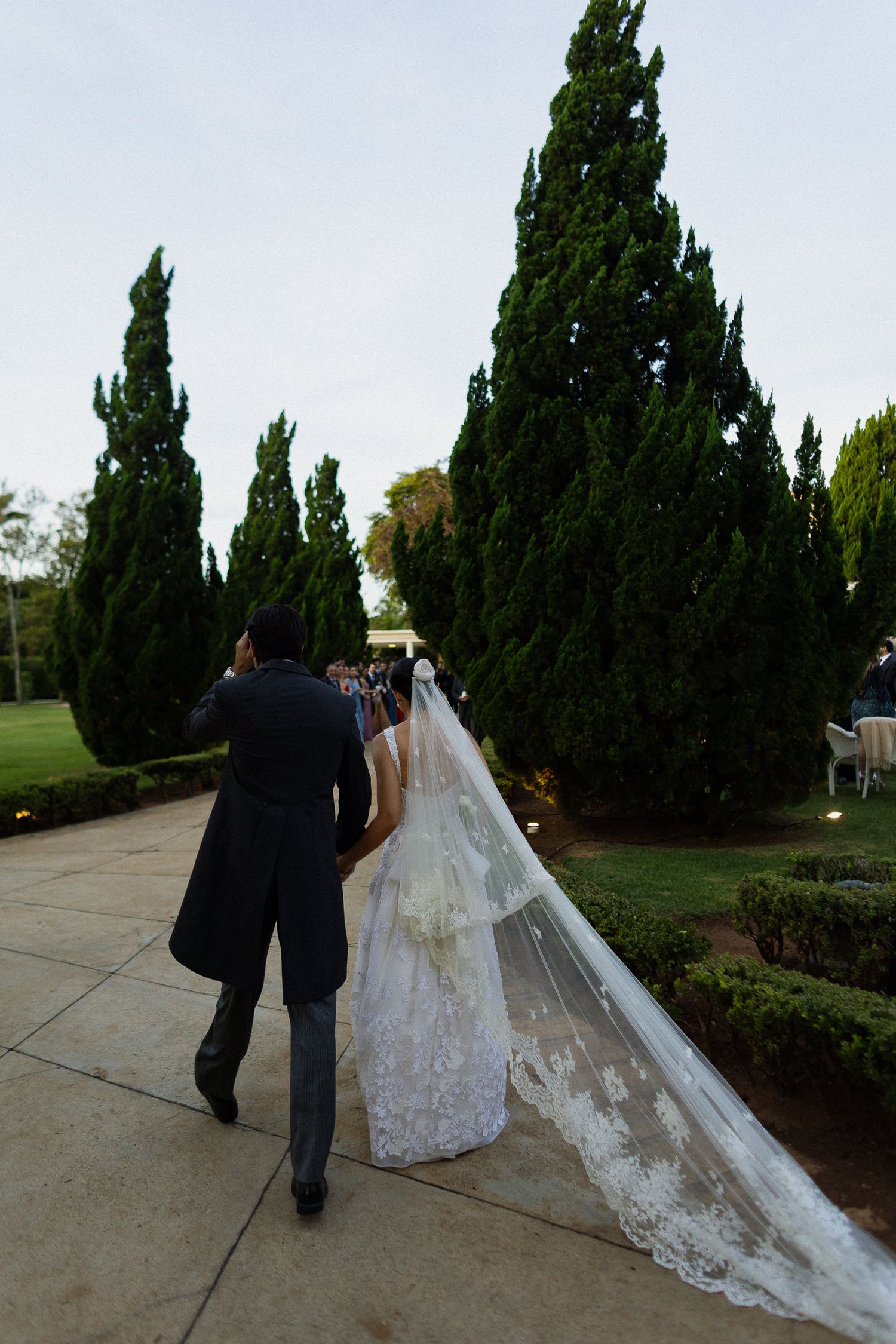 A couple in wedding attire walks along a garden path lined with tall cypress trees on their special day.