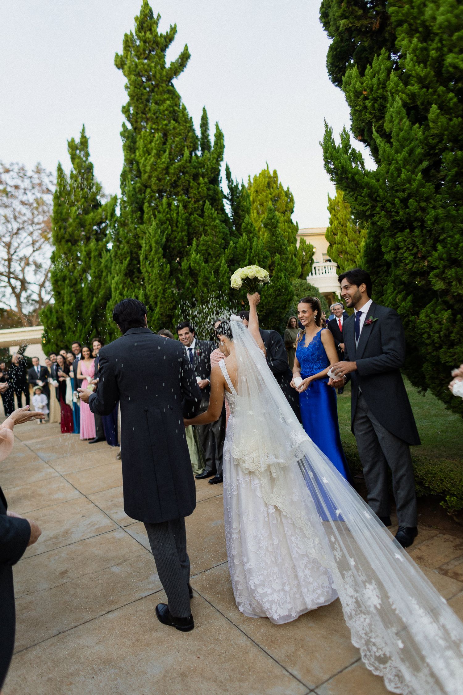 Wedding party gathers on outdoor walkway surrounded by tall cypress trees during sunset ceremony.