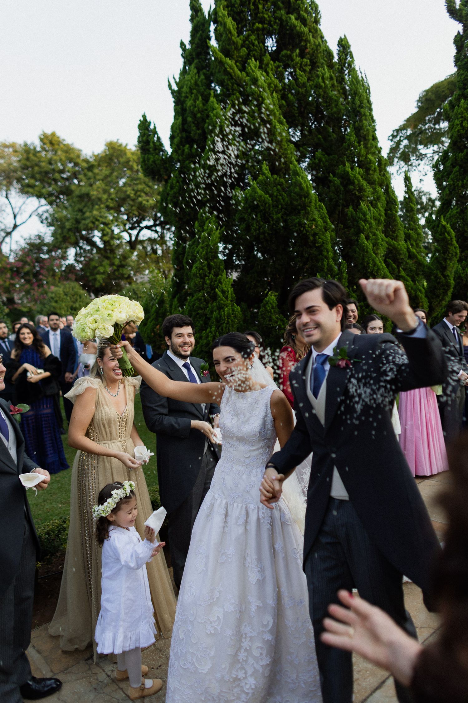 Wedding guests celebrate and dance together outdoors beneath tall cypress trees during a joyful reception celebration.