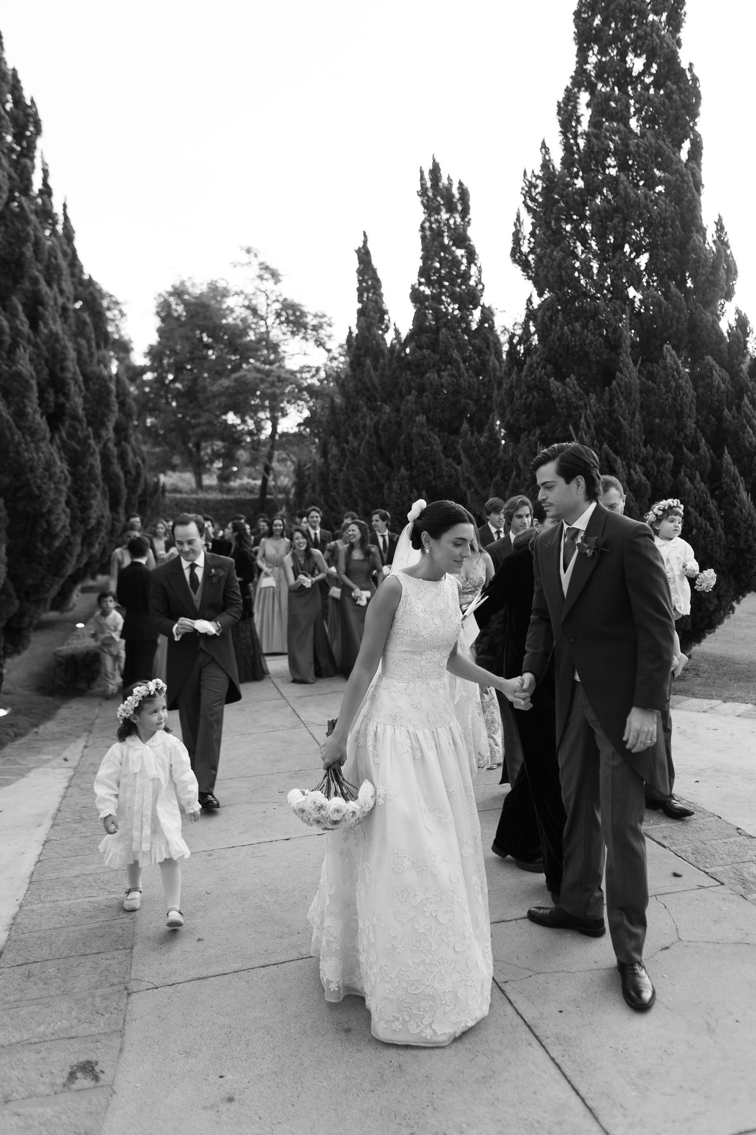 A wedding procession walking along a path lined with tall cypress trees in black and white photography.